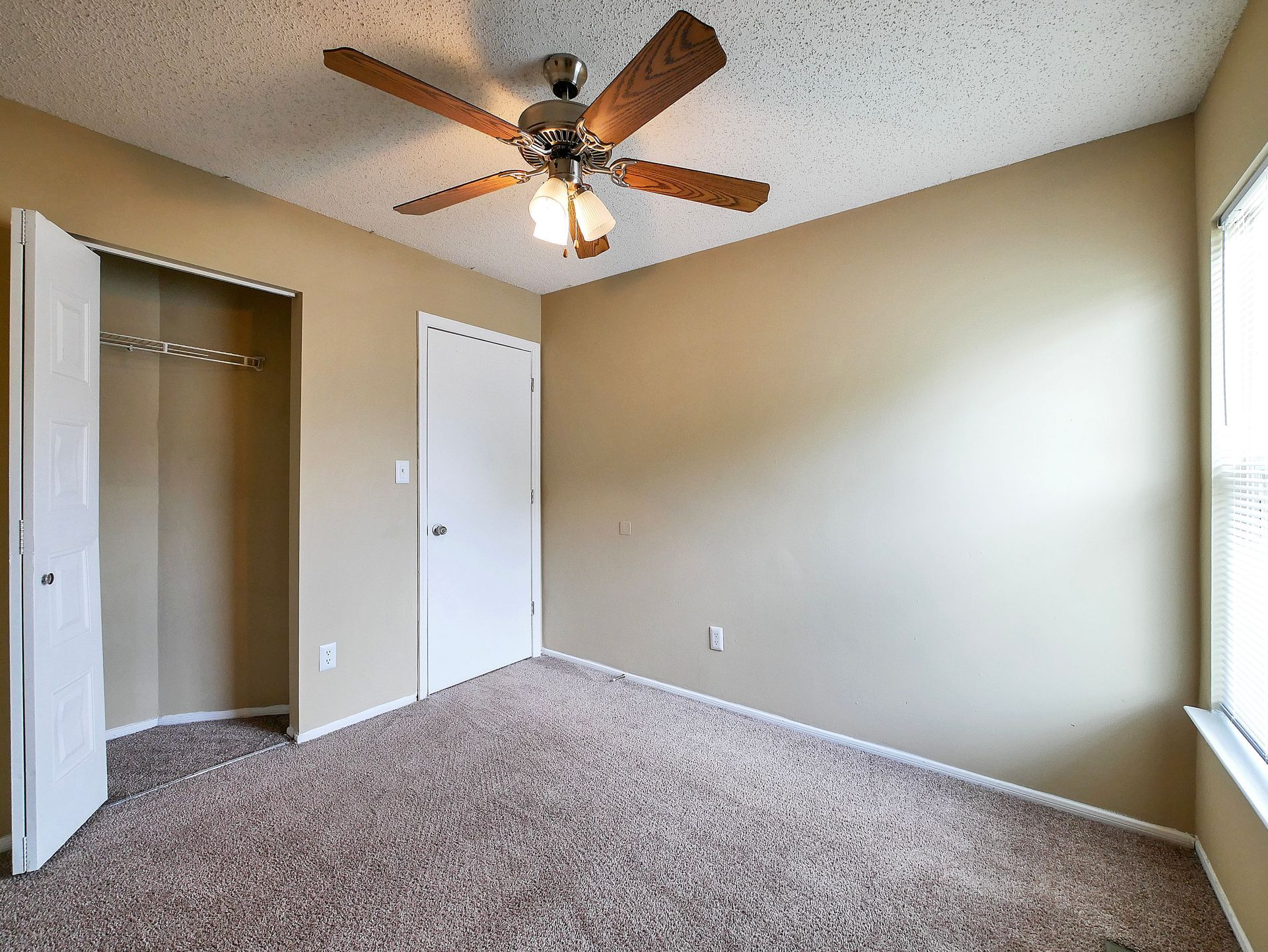 An empty bedroom with tan walls, brown carpet, a white closet door, a ceiling fan, and a window.