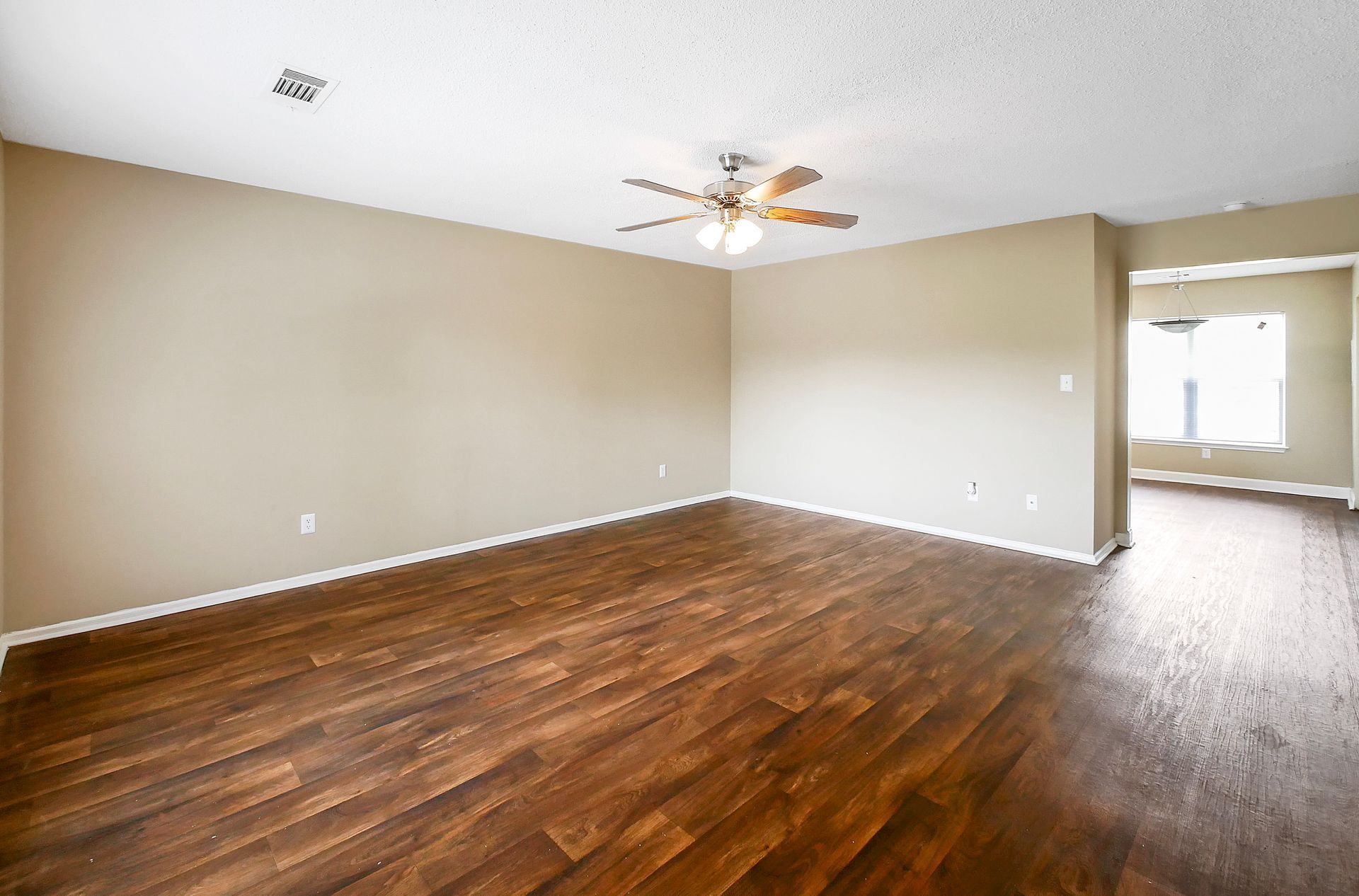 Empty room with warm beige walls, dark wood-look flooring, and a central ceiling fan, leading into an adjacent room.