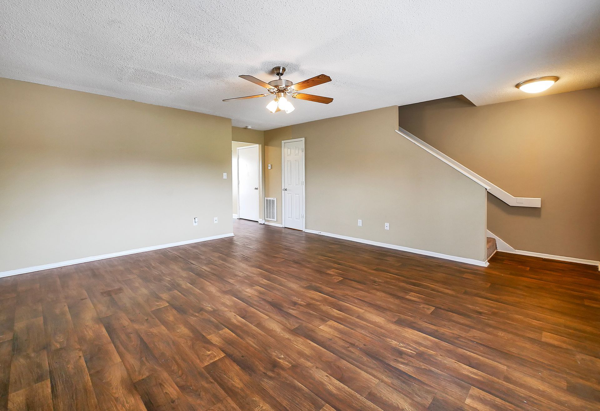 A spacious, empty room featuring tan walls, wood-look flooring, a ceiling fan, and a staircase leading to an upper level.