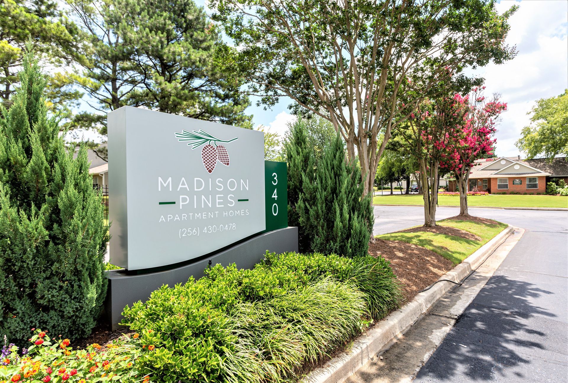 A gray entrance sign for Madison Pines Apartments surrounded by greenery, bushes, and trees on a sunny day.