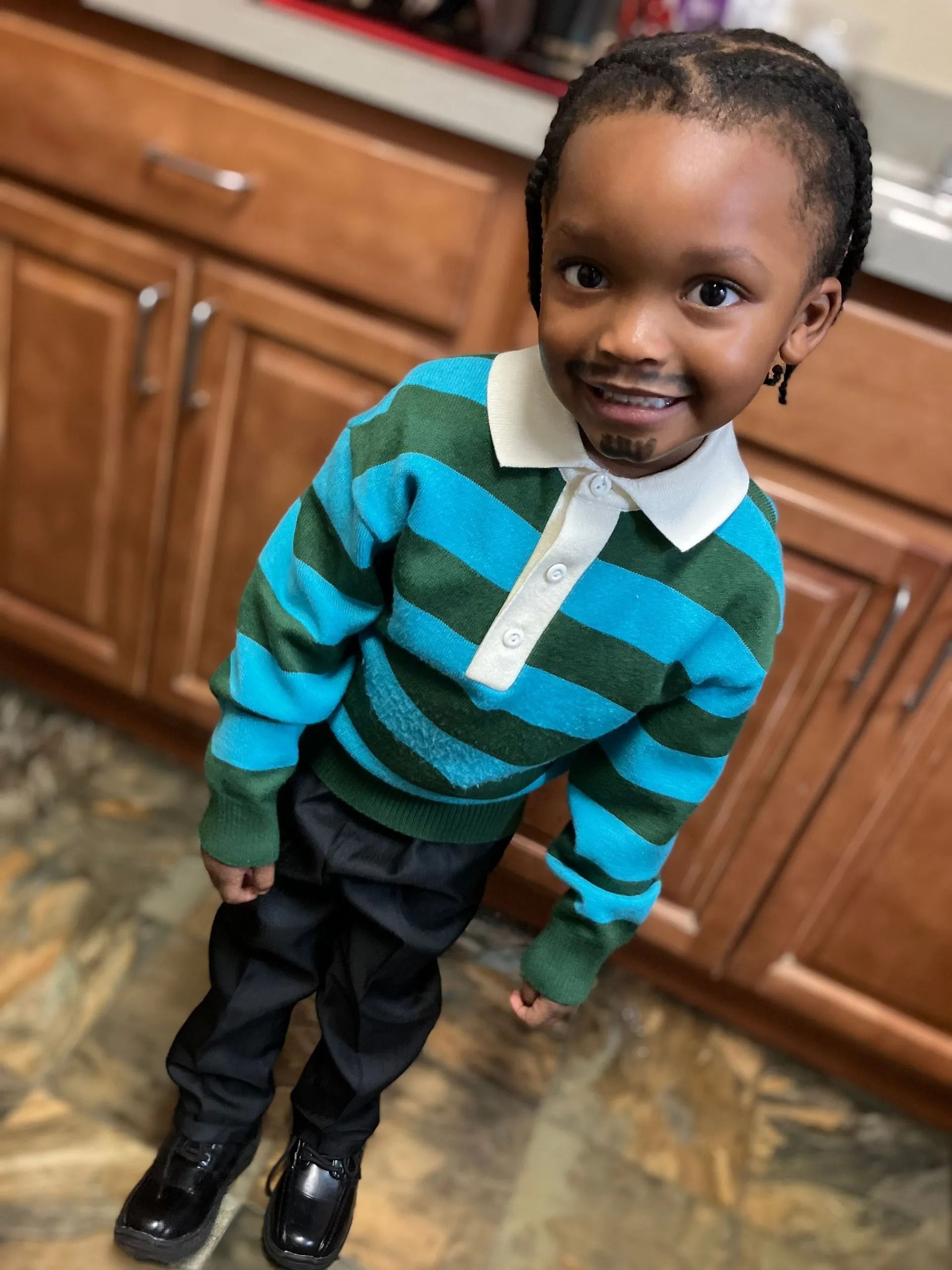 A child wearing a blue and green striped collared sweater, black pants, and shoes stands in a kitchen.