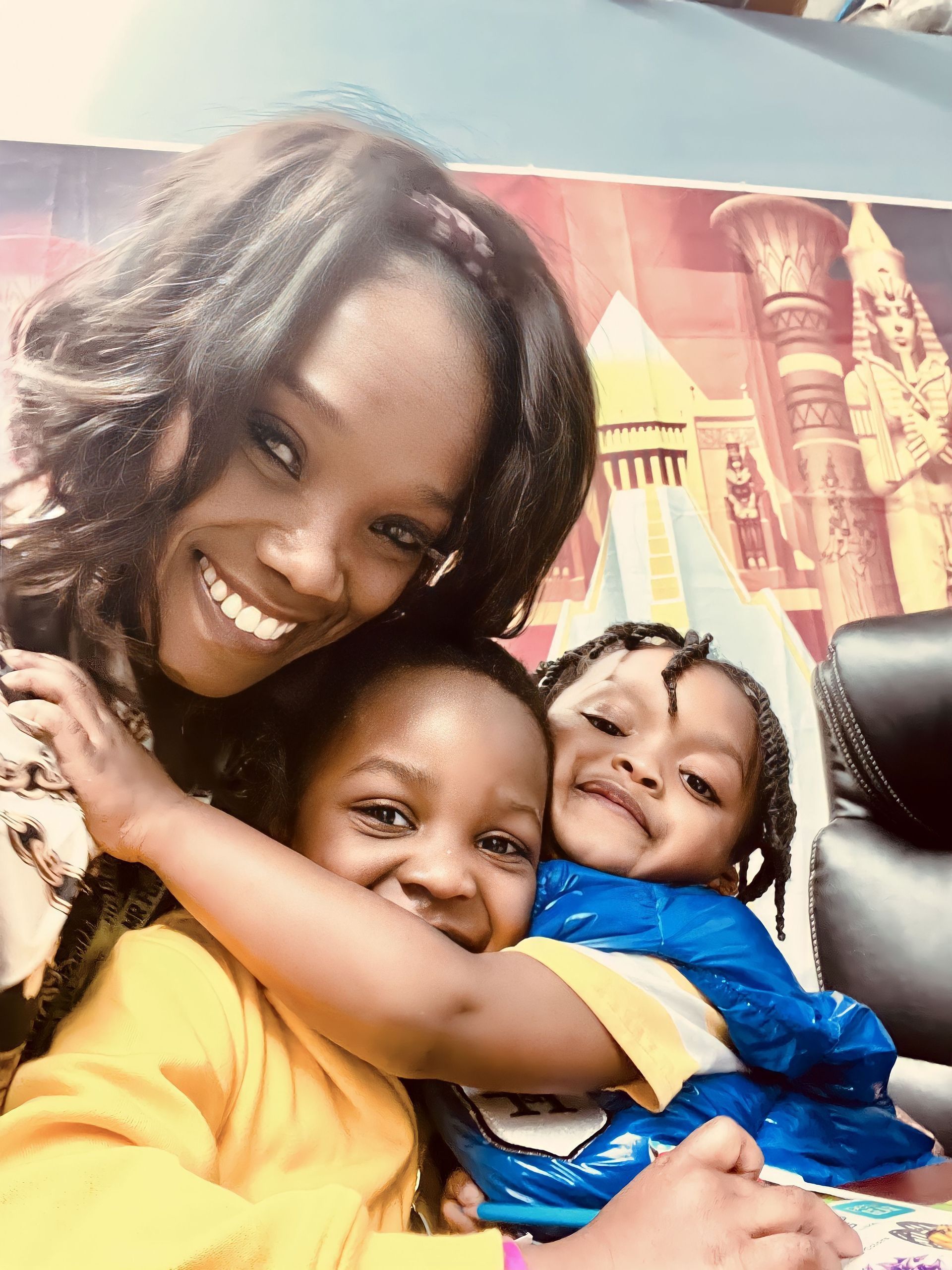 A woman smiling and hugging two children in front of a decorative wall with Egyptian-themed artwork.