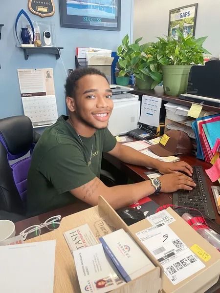 A person in a green shirt sits at a desk typing on a computer in an office setting with plants and office supplies.