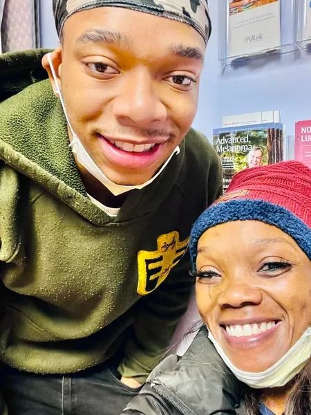Two people smiling for a close-up selfie; one wears an olive hoodie and patterned head covering, the other a red beanie.