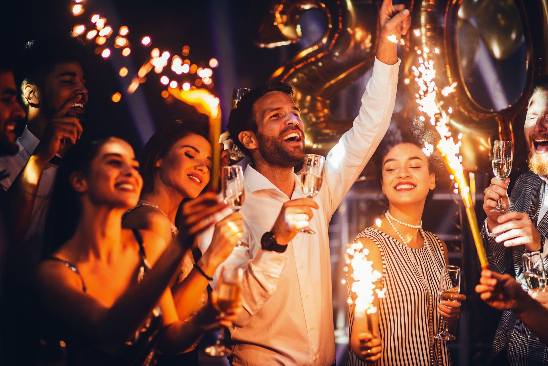 People cheering at a lively night party, holding drinks and sparklers under warm lights