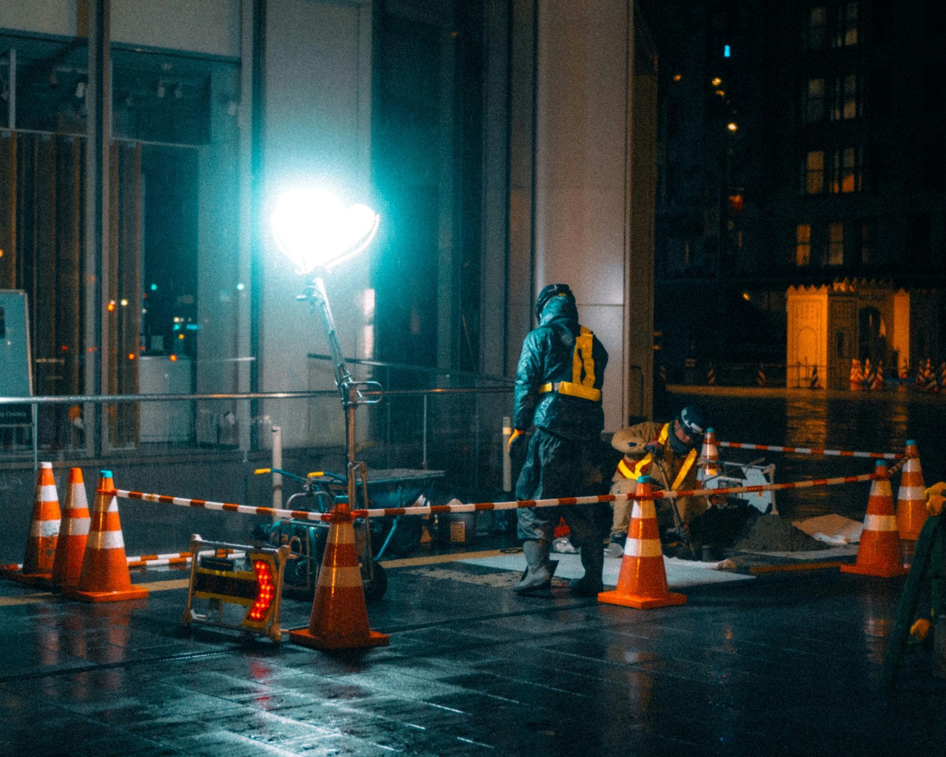 Worker at a nighttime construction site beside orange cones and barriers, lit by a bright floodlight.