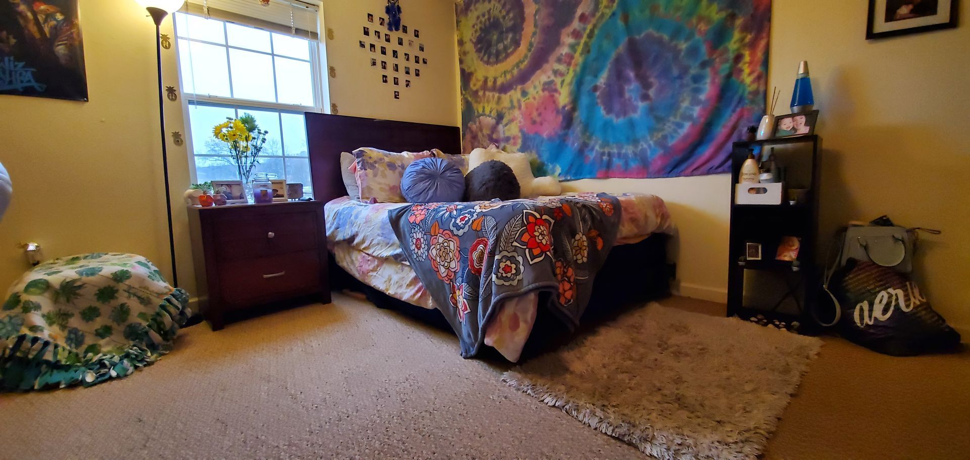 A bedroom with a bed , nightstand , rug and a colorful tapestry on the wall.