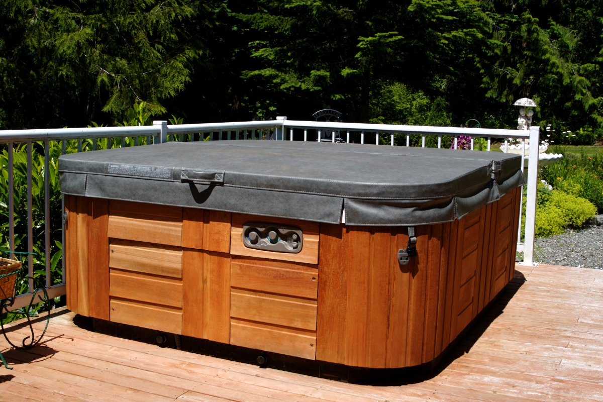 Hot tub with wooden exterior, covered, on a deck with a white railing and trees in the background.