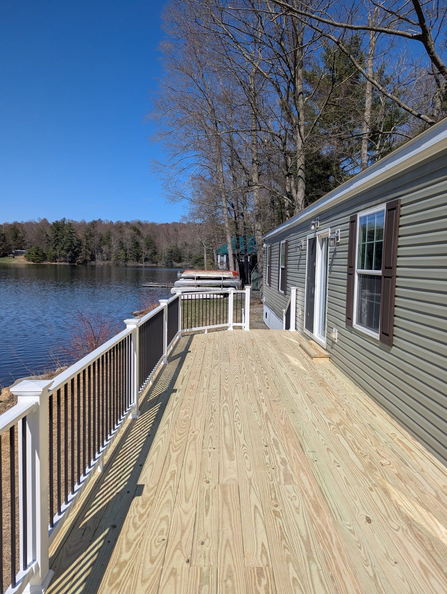 A lakefront home with a wooden deck and railing. Blue sky, water, and trees in the background.