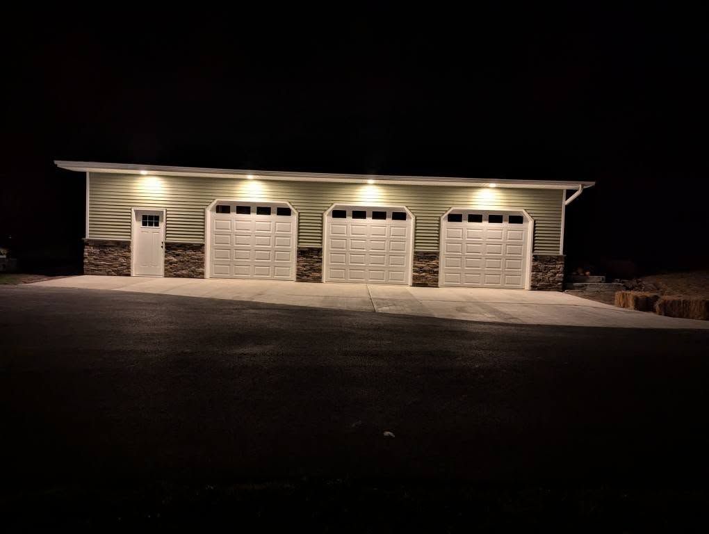 Three-bay garage at night, lit by overhead lights. Stone and light siding, with a door on the left side.