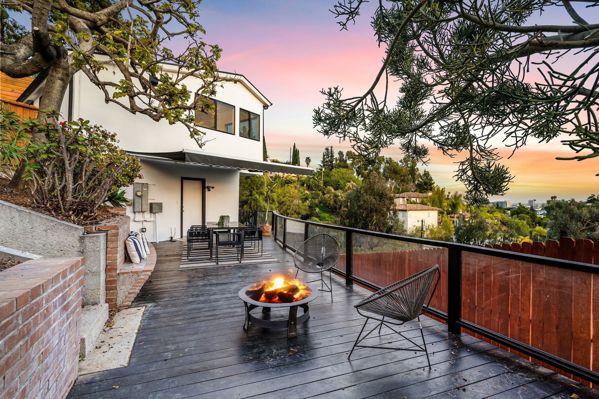 A deck overlooking a landscape with a fire pit, chair, and table set. Sunset colors in the sky.