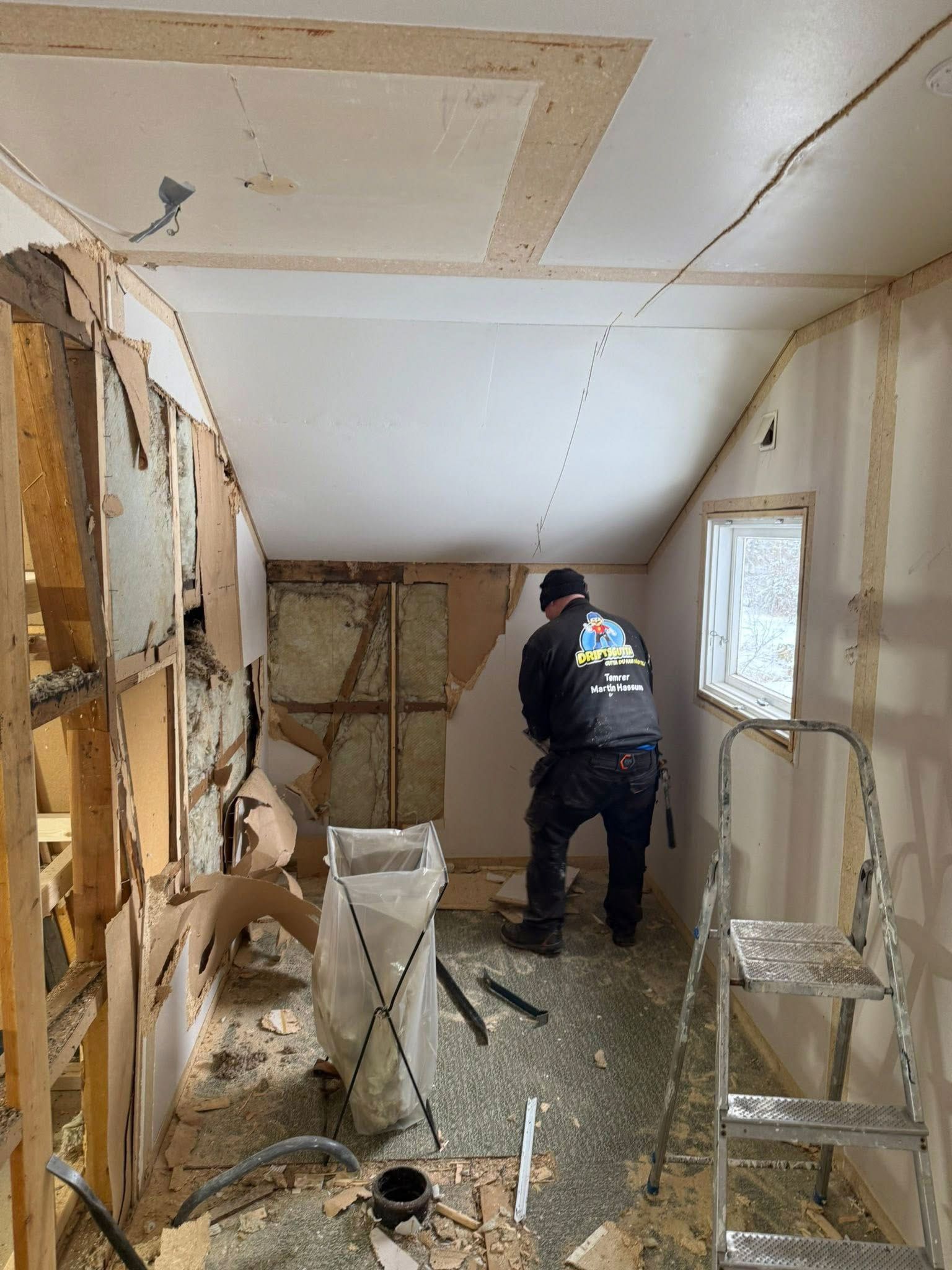 Person som river et rom; eksponert veggkonstruksjon, rusk og en stige tilstede.