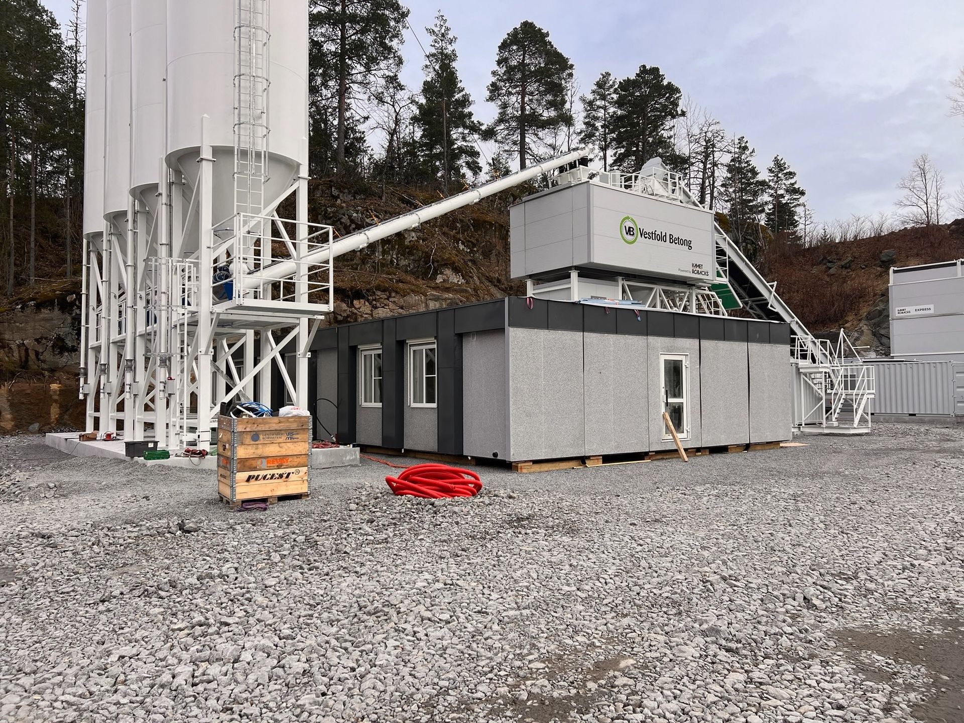 Industrial site with white silos and a gray modular building on a gravel lot in a wooded area.