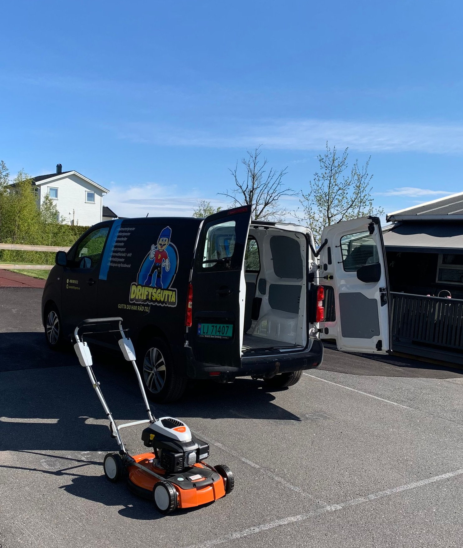 En svart varebil med logo av Driftsgutta, åpne dører og en oransje gressklipper på fortauet.