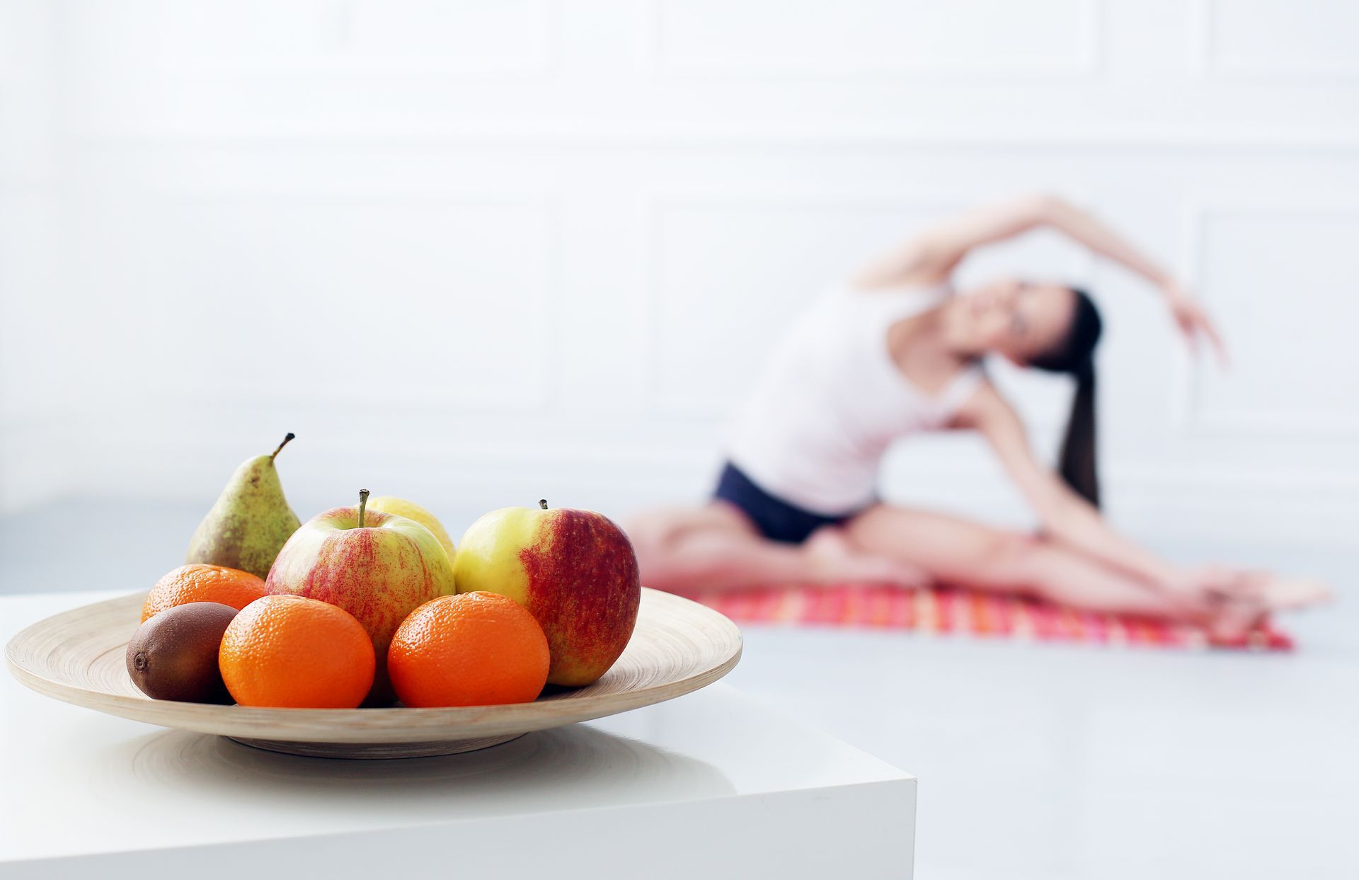 A woman is doing yoga in the background and a bowl of fruit is in the foreground.