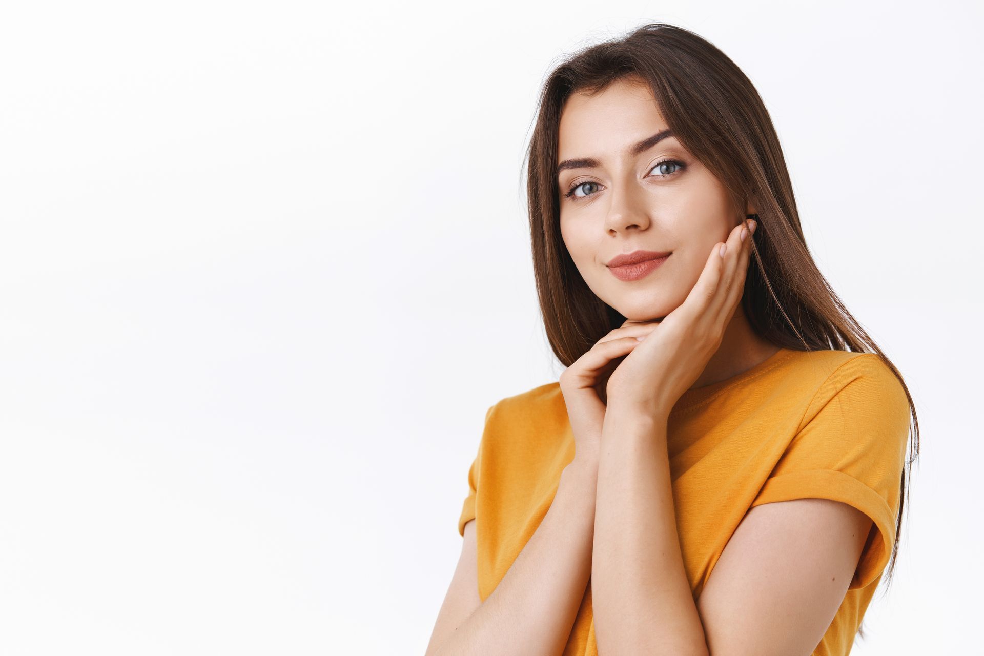 A woman in a yellow shirt is touching her face and smiling.