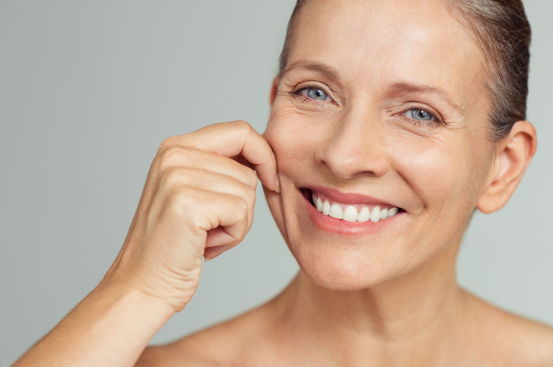 Woman smiling, pulling skin on her cheek, wrinkles visible. Gray background.