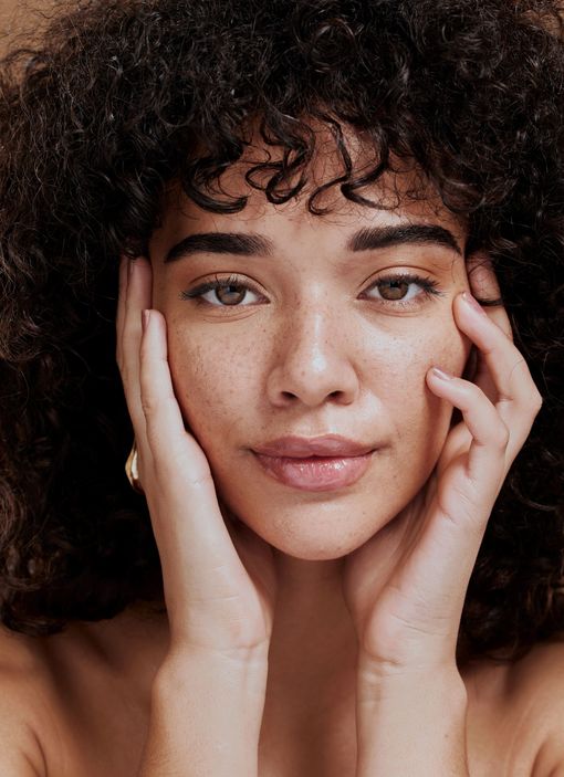 A woman with curly hair and freckles is touching her face with her hands.