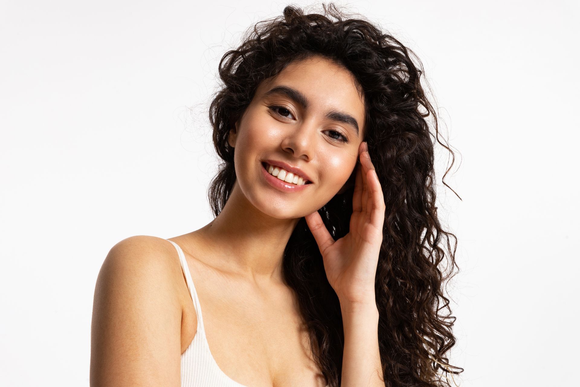 Woman with curly dark hair smiles, touches her temple, wearing a white spaghetti strap top.