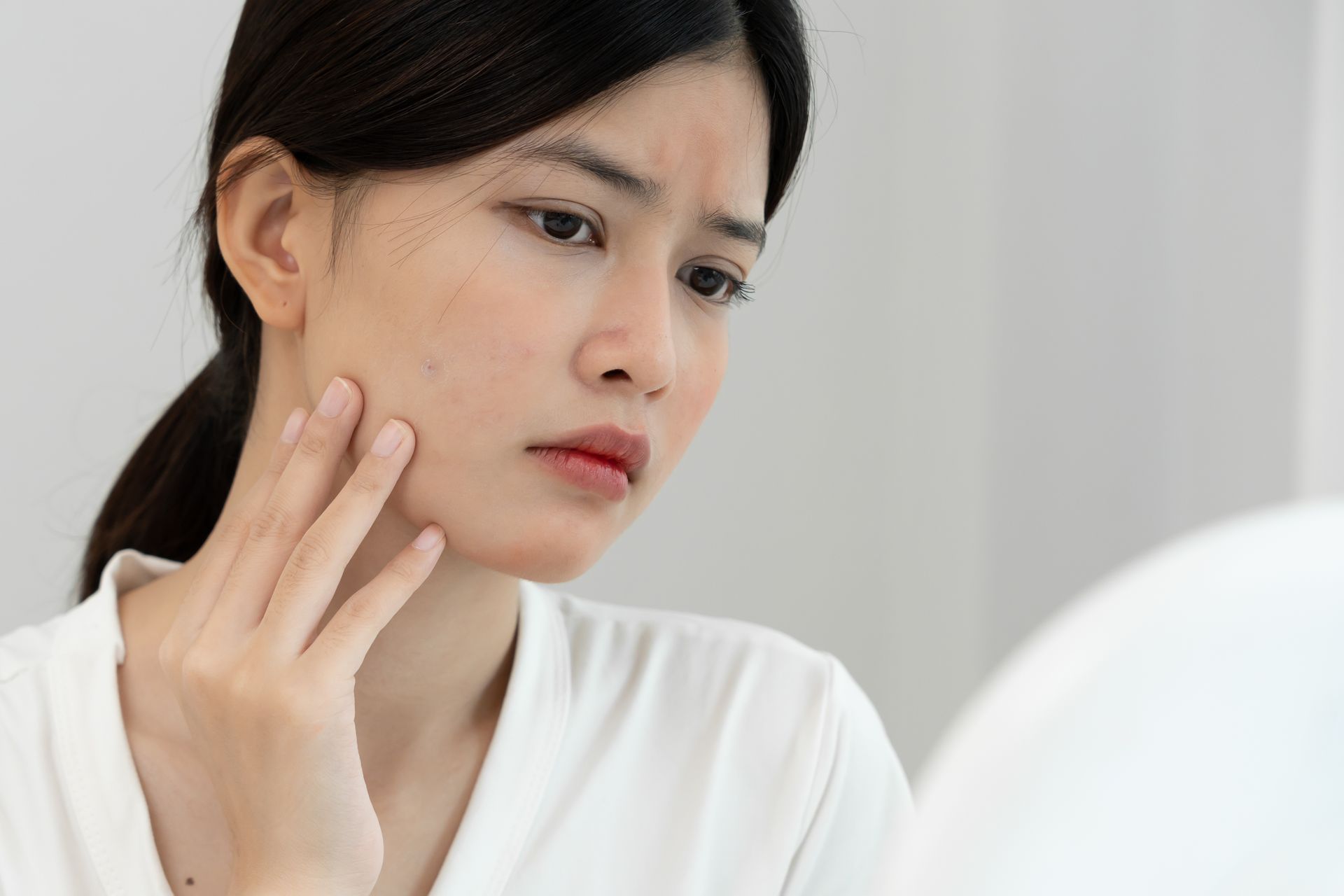 Woman looking at her face in a mirror, touching her cheek with a concerned expression. White background.