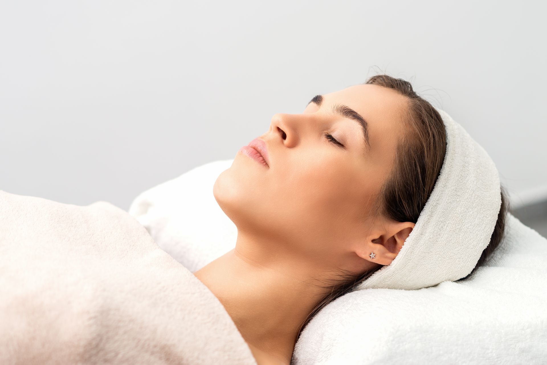 Woman lying down, eyes closed, wearing a headband, on a white bed, wrapped in a towel.