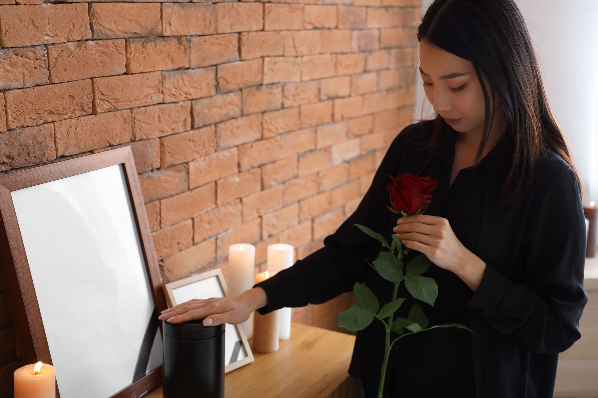 Woman in black holds a red rose, touching a black urn and a picture frame, brick wall in the background.