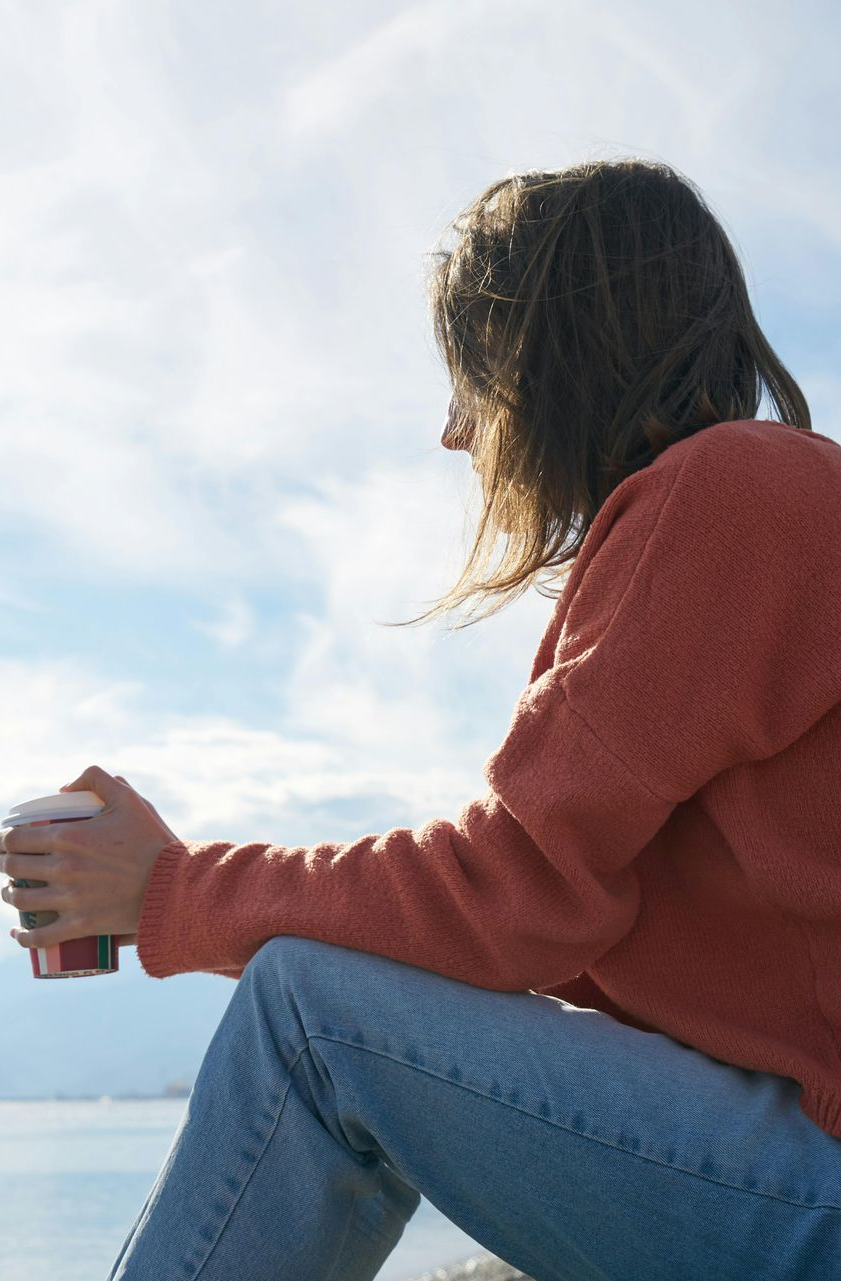 Person in orange sweater and jeans holds a cup, looking at a bright sky over water.
