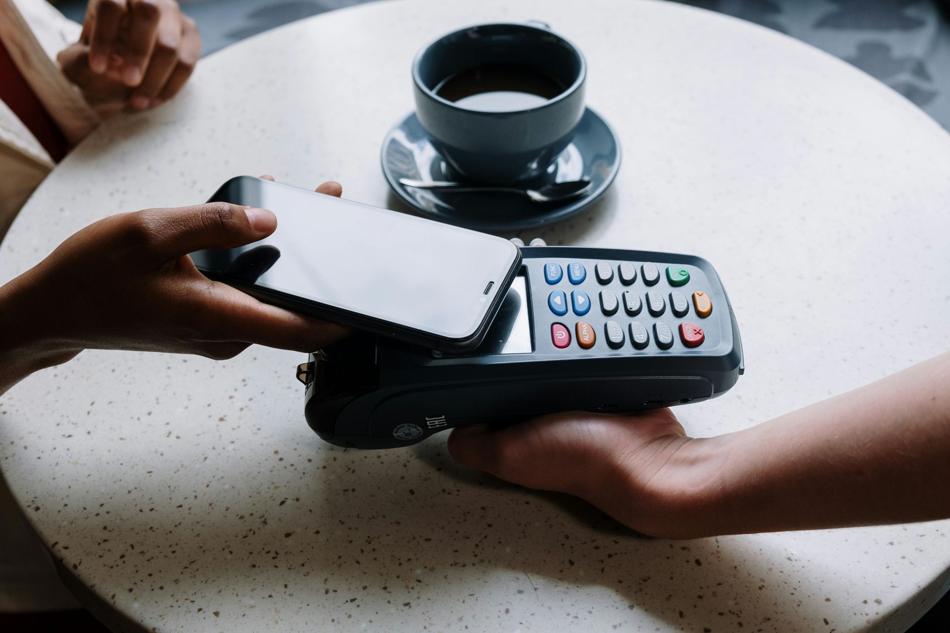 Person using a smartphone to pay at a point-of-sale terminal on a table. Coffee cup in background.
