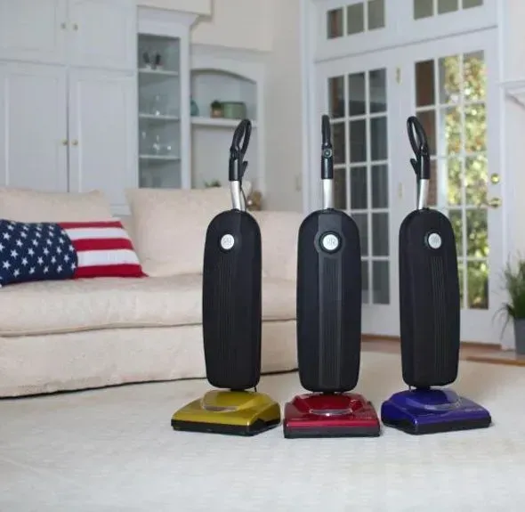 Three upright stick vacuums in yellow, red, and blue on a living room carpet.