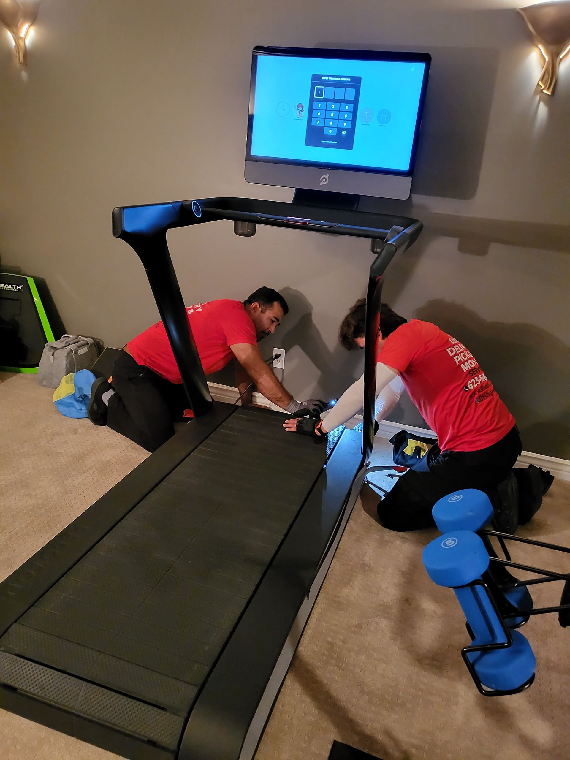 Two people in red shirts are repairing a treadmill in a room with a TV.