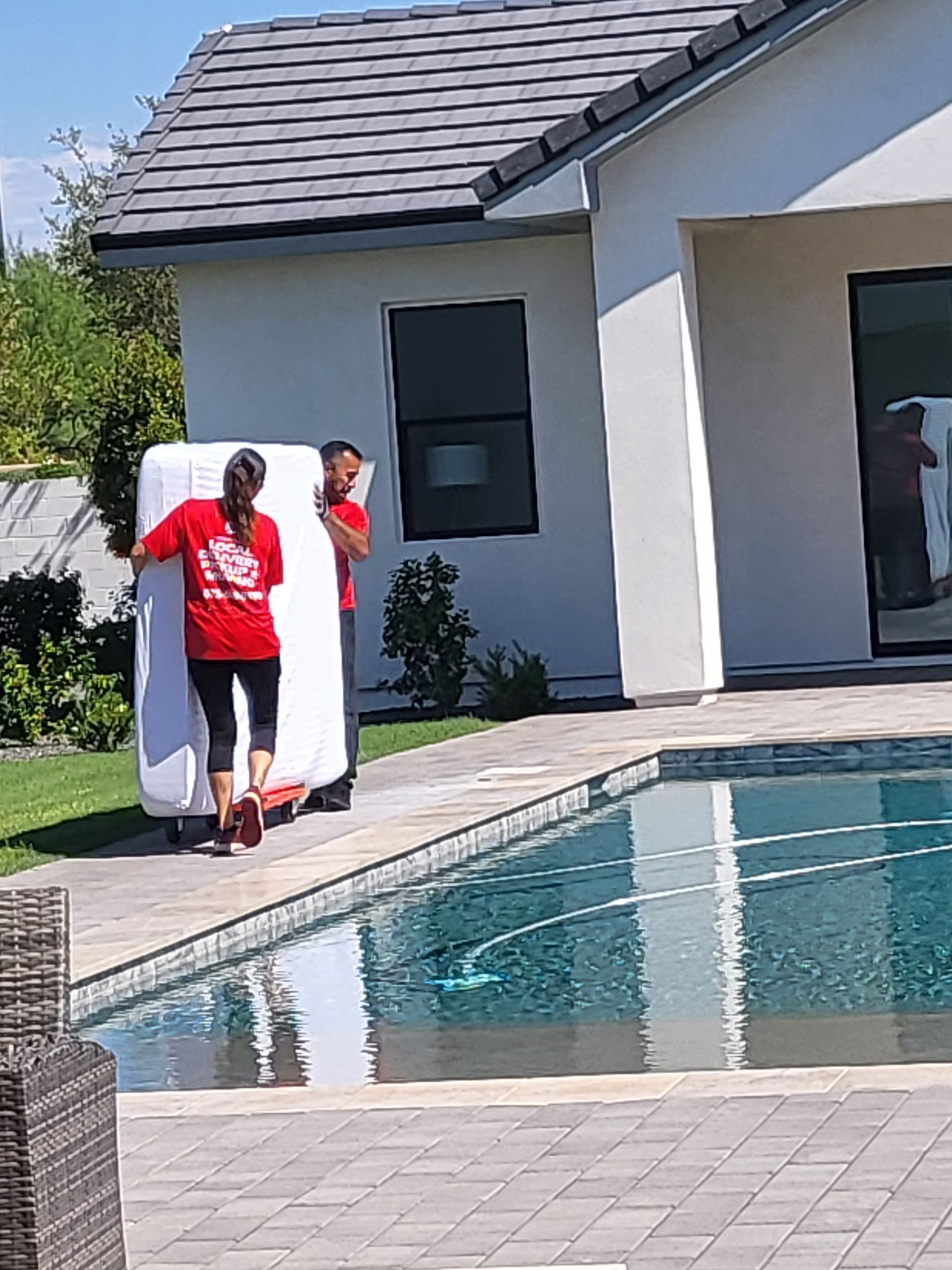 Two people move a wrapped object near a pool. A house and landscaping are in the background.