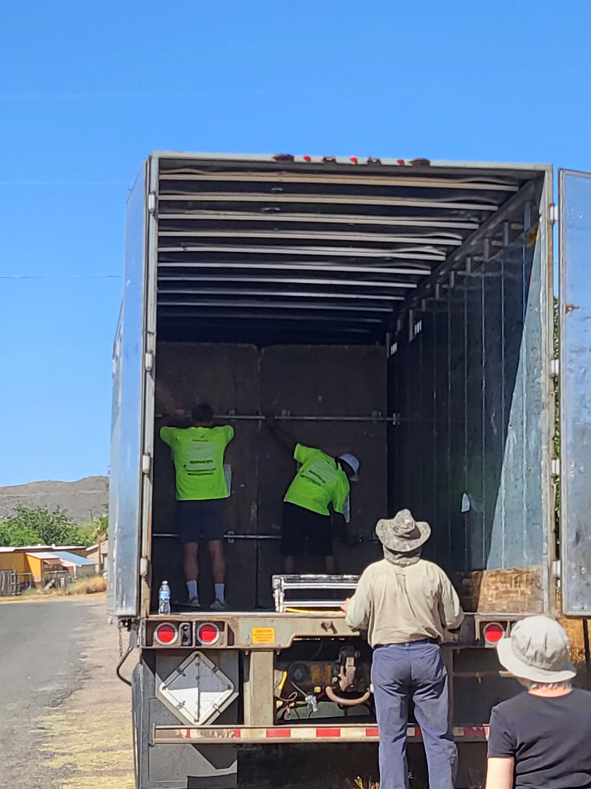 Three people loading items into a truck trailer. Two inside, wearing green shirts, and one outside, wearing a hat, observing.