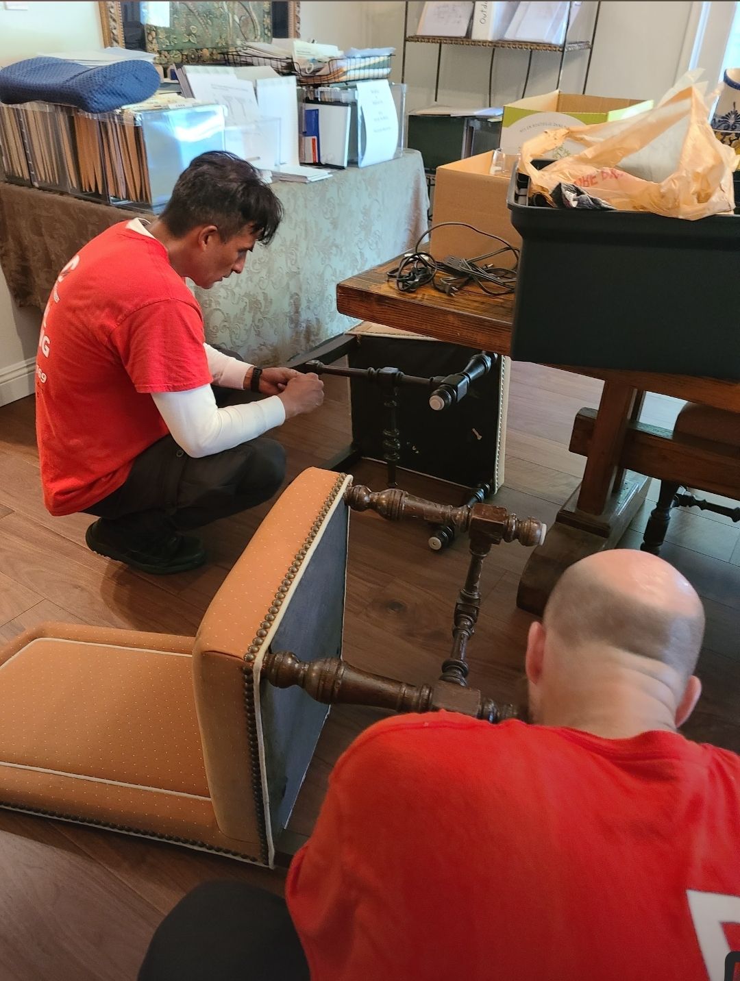 Two people in red shirts assembling furniture indoors. One kneels, the other is seated. Brown wooden floor.