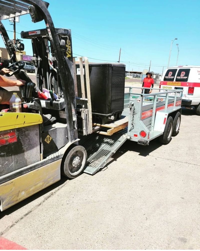 Forklift loading an AC unit onto a trailer in a parking lot on a sunny day. A person in a red shirt stands nearby.
