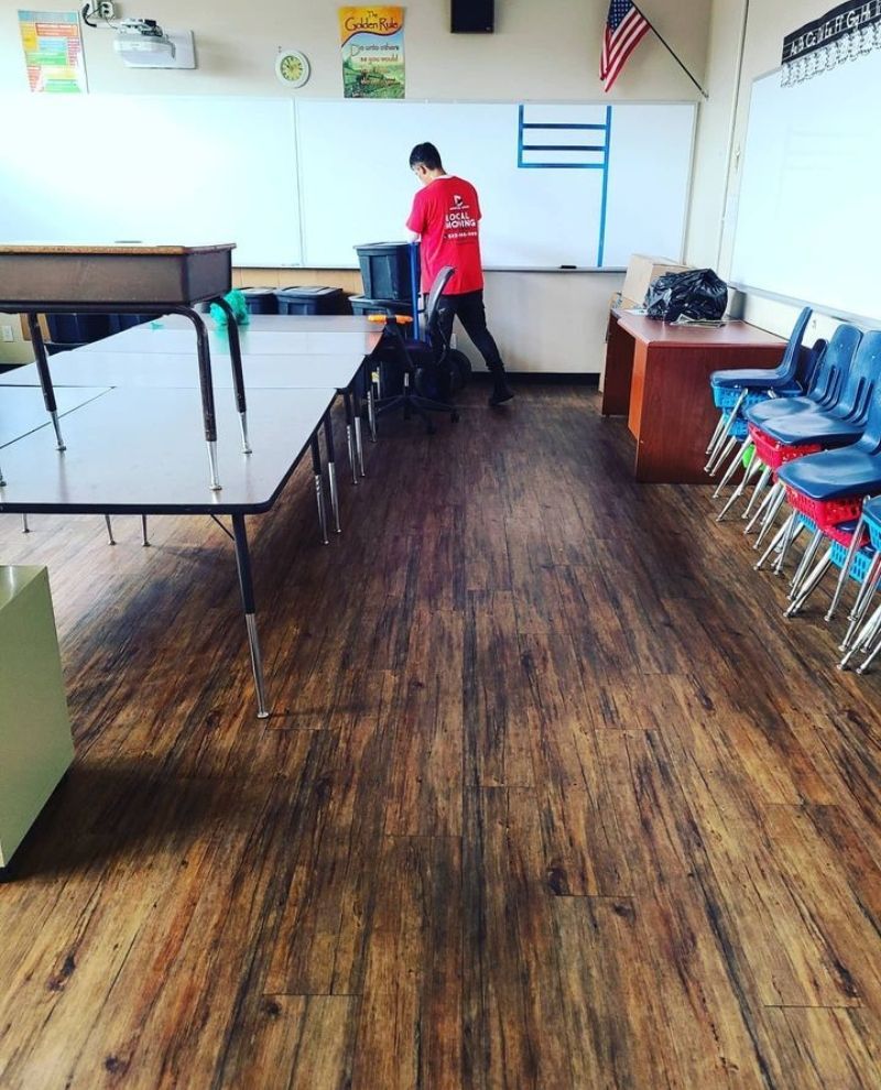 A person in a red shirt cleans a classroom with wood-look flooring. Tables, chairs, and a whiteboard are present.