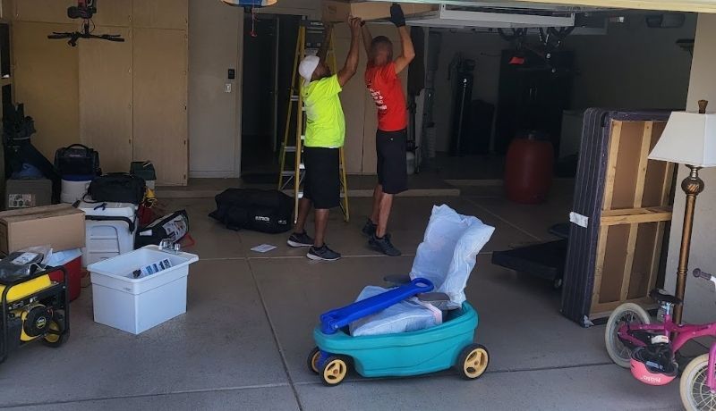 Two people lifting a box inside a cluttered garage, a child's toy car in the foreground.