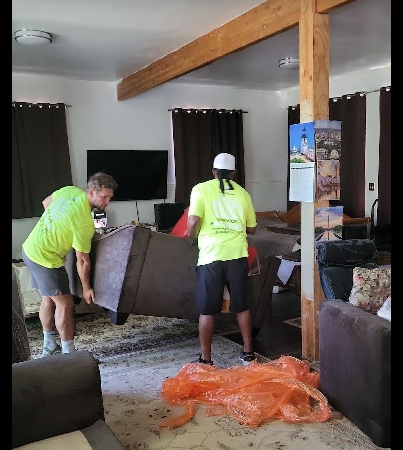 Two movers in neon shirts carry a sofa in a living room; orange wrapping on the floor.