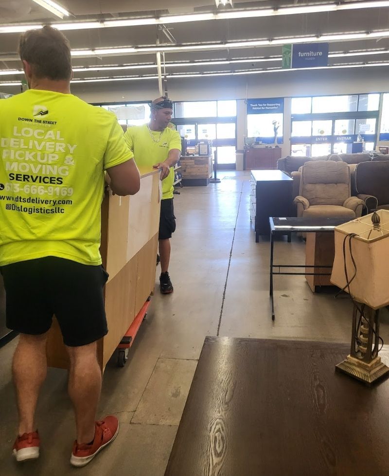 Two men in yellow shirts move a large box on a dolly through a store with furniture.