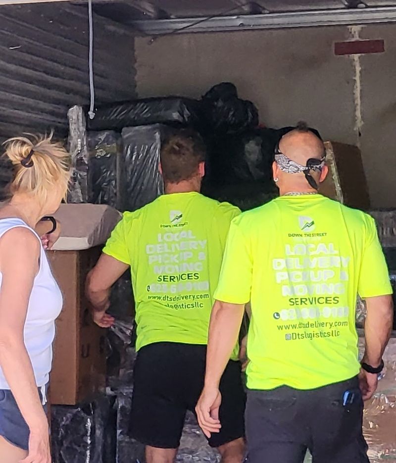 Movers loading a truck with wrapped furniture and boxes. A woman watches them inside the truck.