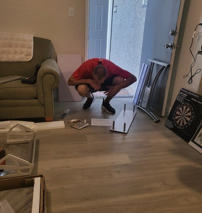 Man crouching, assembling furniture indoors; door, dartboard, boxes visible.