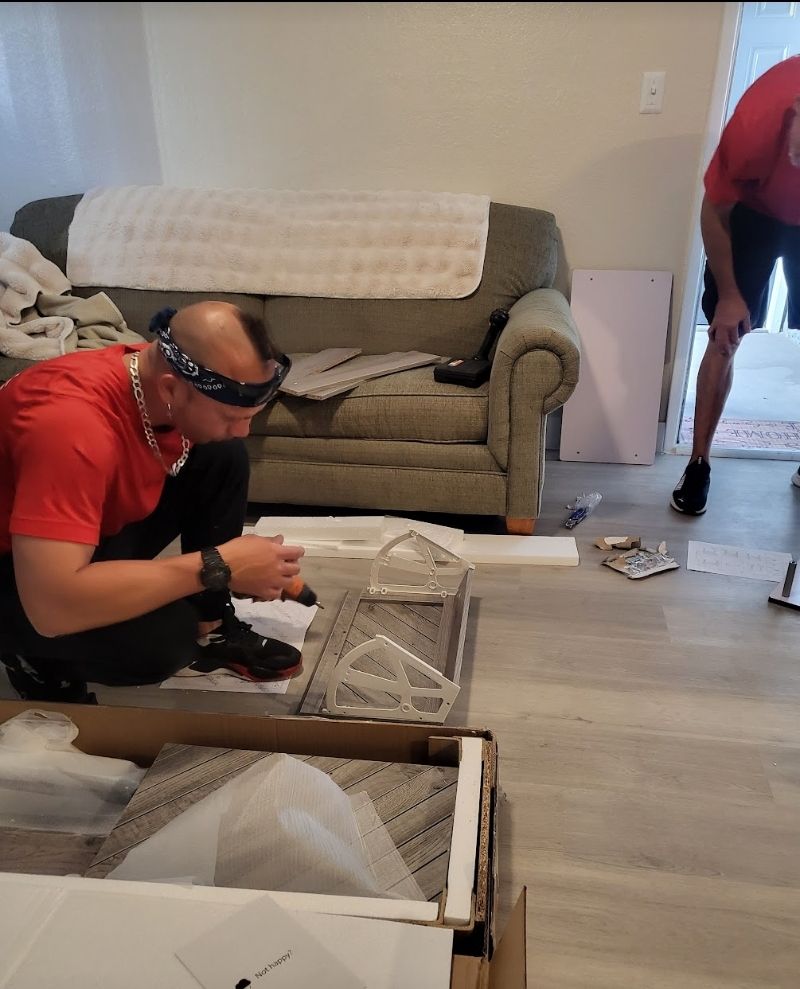 Man assembling furniture on floor, another man stands nearby. Indoors, gray flooring, green sofa in the background.