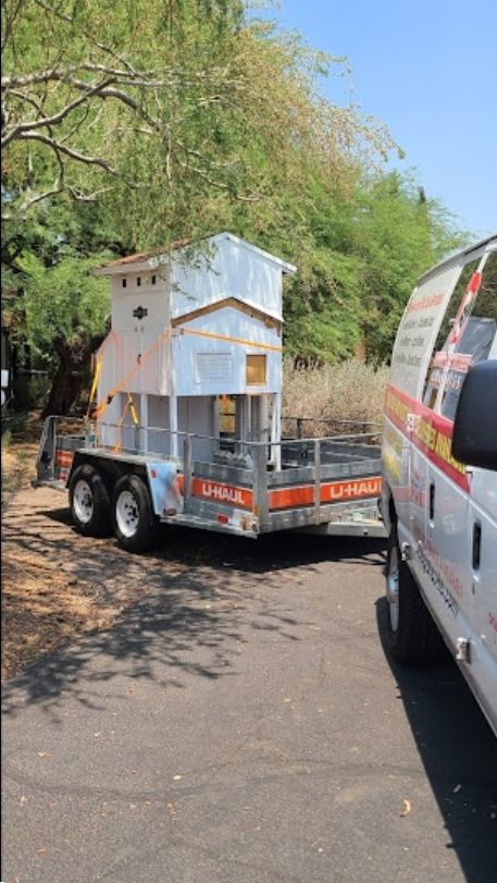 A small, weathered wooden structure on a trailer, next to a service van, outdoors.