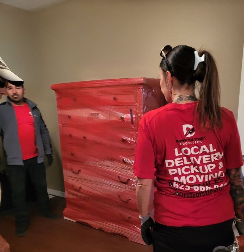Two people moving a red dresser wrapped in plastic; indoors.