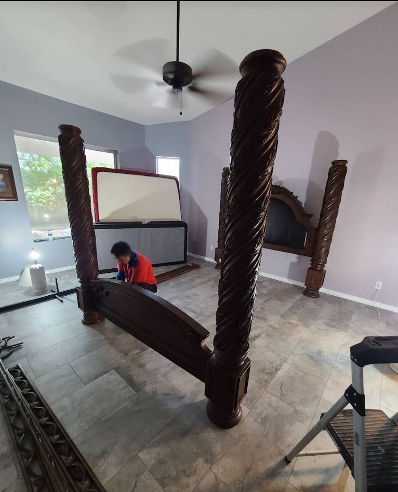 A person assembling a dark wood bed frame with tall, spiral posts in a bedroom with gray tile floors.
