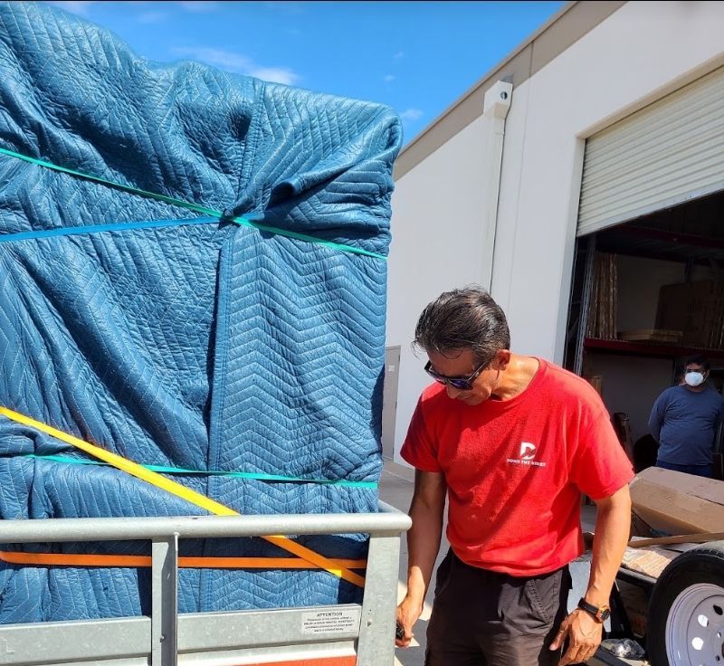 Man in red shirt inspects furniture secured on a trailer with blue moving blankets, near a loading dock.