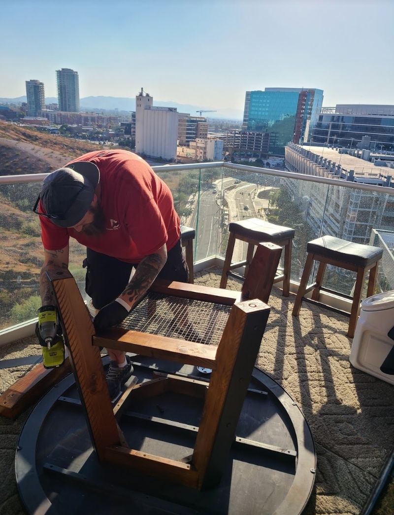 Person cutting wood on a deck overlooking a city skyline. Brown chairs and a fire pit visible. Sunny day.
