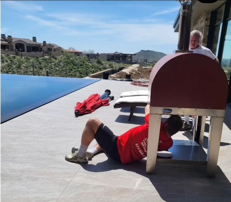 Man in red shirt adjusts a metal cabinet on a patio near a pool, another person looks on.