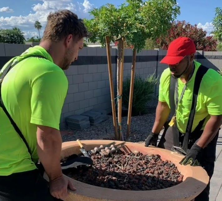 Two workers in green shirts carry a large planter filled with rocks.