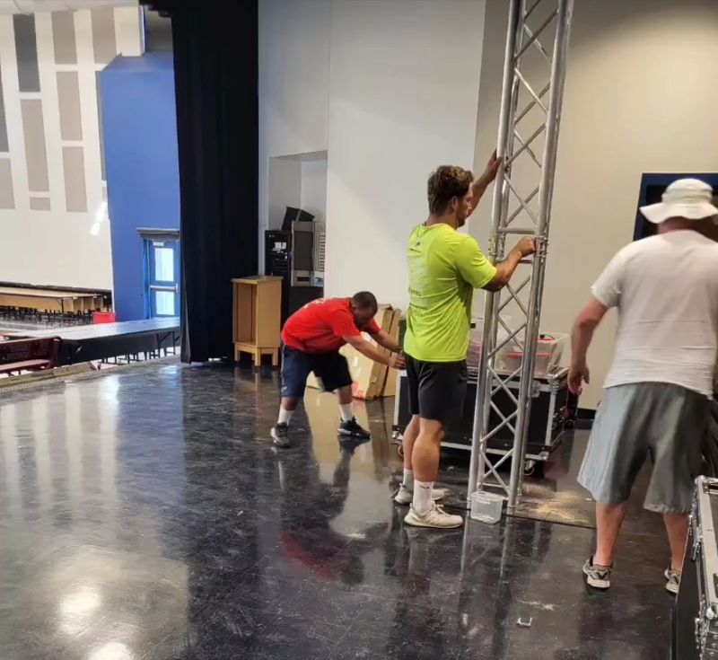 Three people lift a metal truss in a theater. Two wear shorts; one, a lime green shirt. Empty stage.