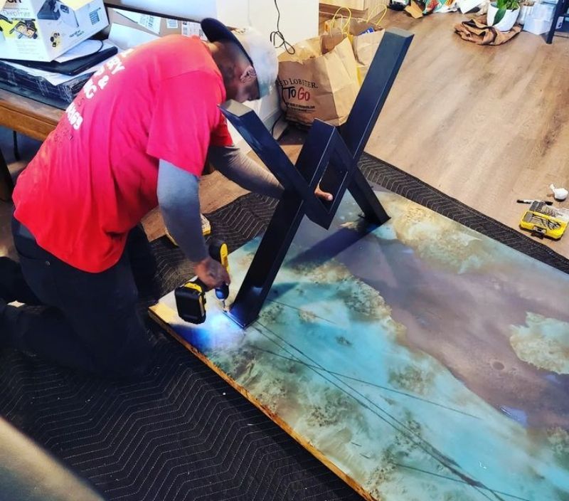 Person attaching black table legs to a teal and brown epoxy resin table top with a power drill.
